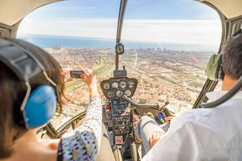 Panoramic aerial view of Barcelona city and coastline from a helicopter cockpit, with a passenger capturing the stunning landscape with their phone, enjoying a unique flight experience.