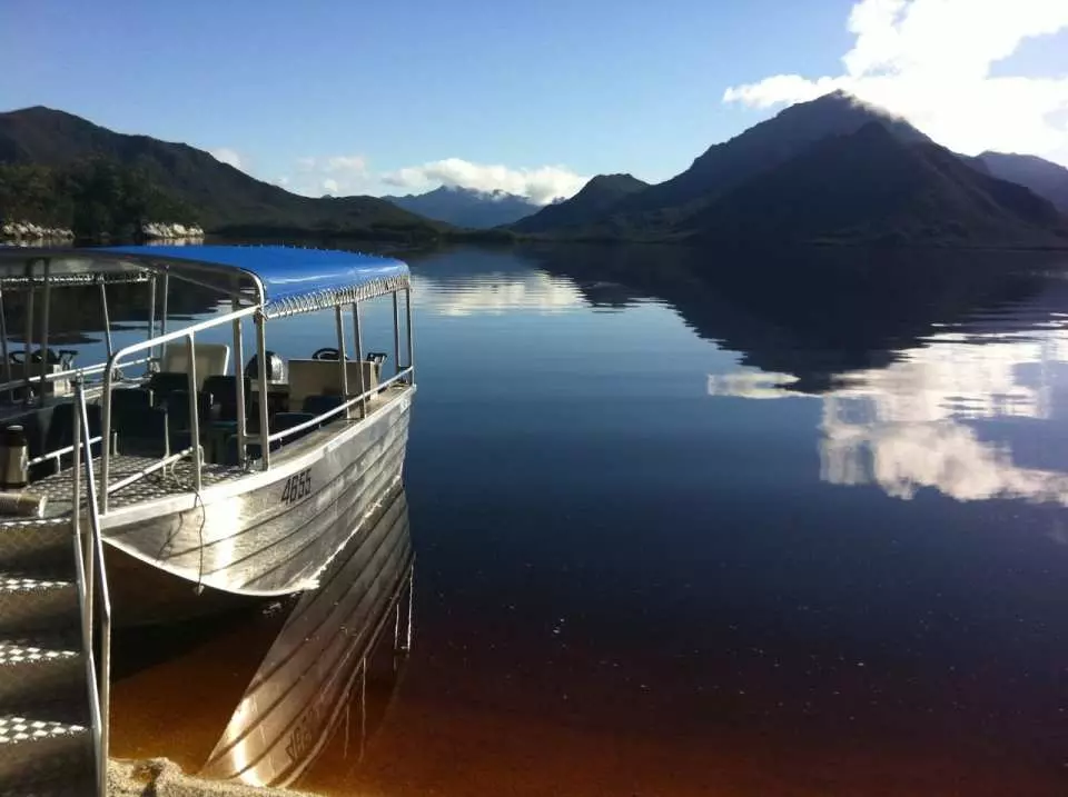 A scenic boat docks by the calm shore of Tasmania's Bathurst Harbour, with clear reflections of distant mountains, ready to embark on an exploration of the southwestern wilderness waters.