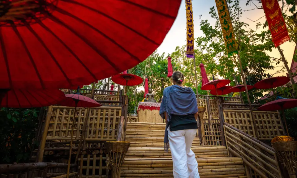 A visitor walks along a bamboo pathway in Chiang Mai Old City, heading towards a Buddha statue surrounded by red parasols, experiencing the unique tranquility and natural ambiance of a Thai temple walking tour.