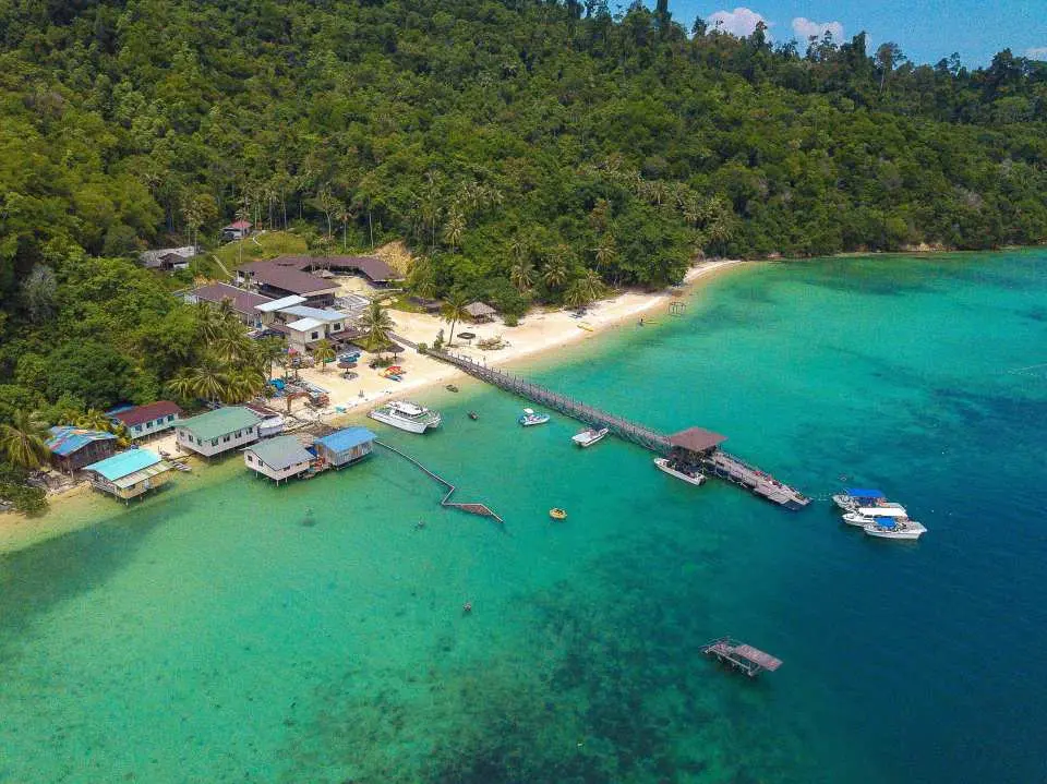 Aerial view of Sabah's Dreamer Island day tour, featuring a lush green island, white sand beach, and a pier extending into crystal blue waters, ideal for diving and island relaxation.