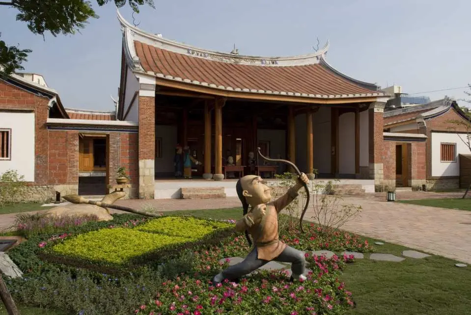 A vibrant courtyard scene at Fengyi Academy with a lively statue of a child archer amidst green landscaping and flowers, offering an engaging historical and interactive experience for visitors.