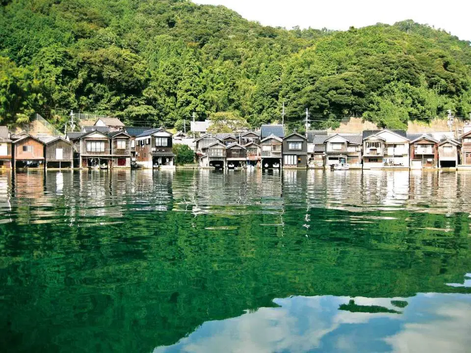 京都伊根湾の岸辺に並ぶ伝統的な木造舟屋が、澄んだエメラルドグリーンの水面に鮮明に映り込み、背景の青々とした山林と絵のように美しい景色を織りなす。