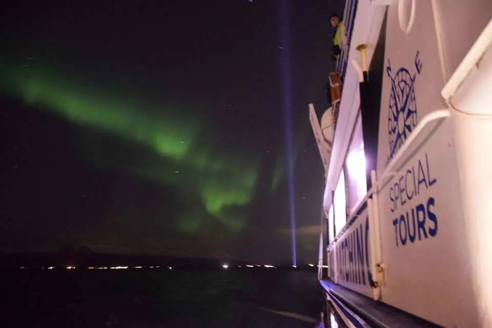 冰島雷克雅維克極光船在夜間航行，船身旁的海面映照著遠方城市燈光，頭頂上則是舞動的綠色極光，提供難忘的體驗。