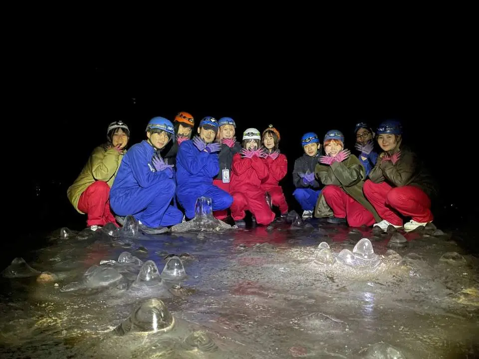 A group of tourists in protective suits and helmets posing on an icy surface inside a lava cave in Aokigahara, with headlamps illuminating their faces, enjoying a unique ice cave adventure.