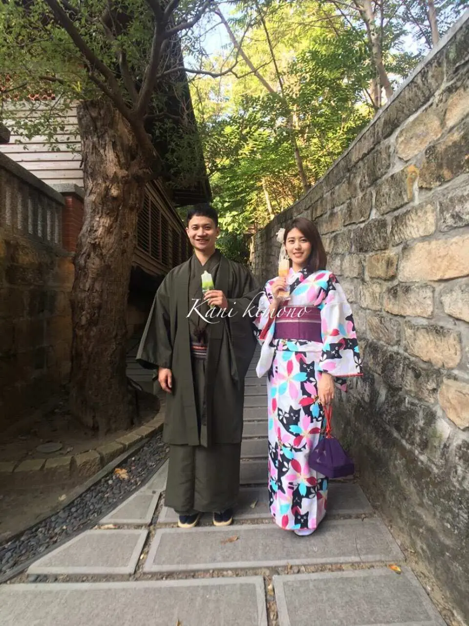 一對情侶在北投充滿綠意的石板小徑上漫步，男生穿墨綠和服，女生穿多彩浴衣，體驗穿梭日式園區的樂趣。