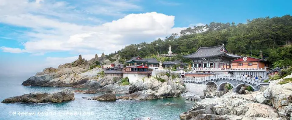 釜山海東龍宮寺の広角パノラマ。壮麗な寺院群、石のアーチ橋、ごつごつした海岸線が見事に融合し、紺碧の海面が白い雲と青空を映し出しています。