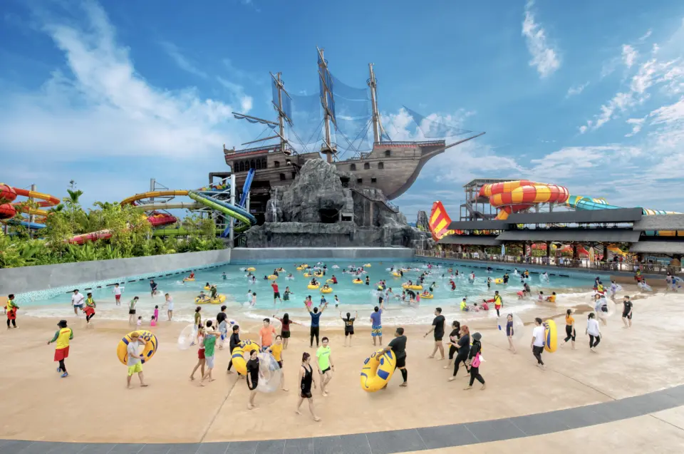 Expansive wave pool at SplashMania Water Park, with many visitors enjoying the water, featuring a large pirate ship themed structure and multiple thrilling water slides in the background.