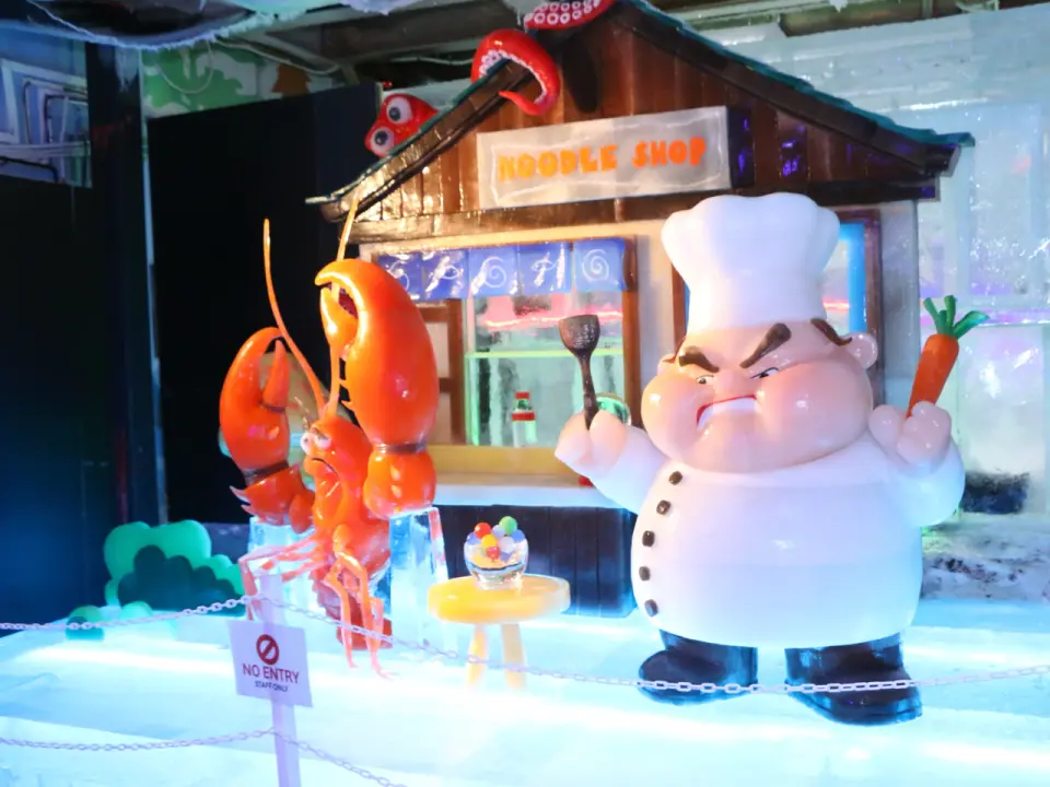 A creative ice sculpture scene inside Singapore Snow City features an ice 'Noodle Shop' facade, a chef figure, and a giant lobster ice sculpture, adding a touch of fun.