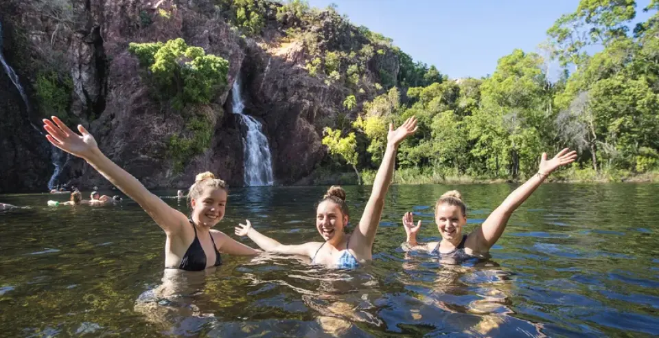 Three joyful women swim in the crystal-clear natural pools of Litchfield National Park, with a stunning waterfall and lush tropical rainforest in the background, enjoying a refreshing moment.