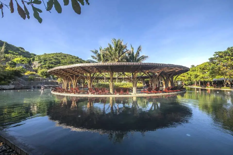 The elegant circular gazebo over water at Hon Tam Island Mud Bath Resort, Nha Trang, adorned with palm trees under sunlight, with its reflection perfectly mirrored on the calm water surface.