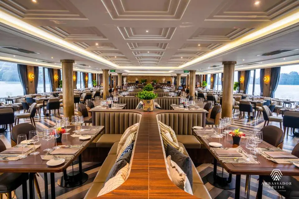 Spacious and elegantly decorated main dining hall on the Ambassador luxury cruise, with tables set for fine dining and panoramic views of Halong Bay.