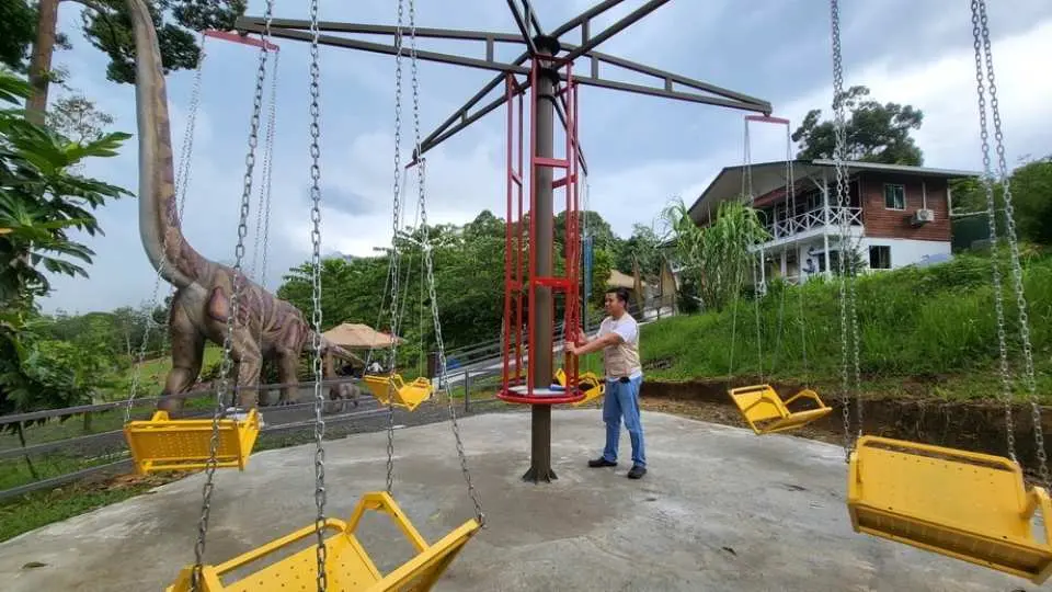 A visitor operating a vibrant yellow chain carousel ride for children, with a giant Brachiosaurus replica in the background, amidst the family-friendly attractions at Jurassic Land Kiulu, Sabah.
