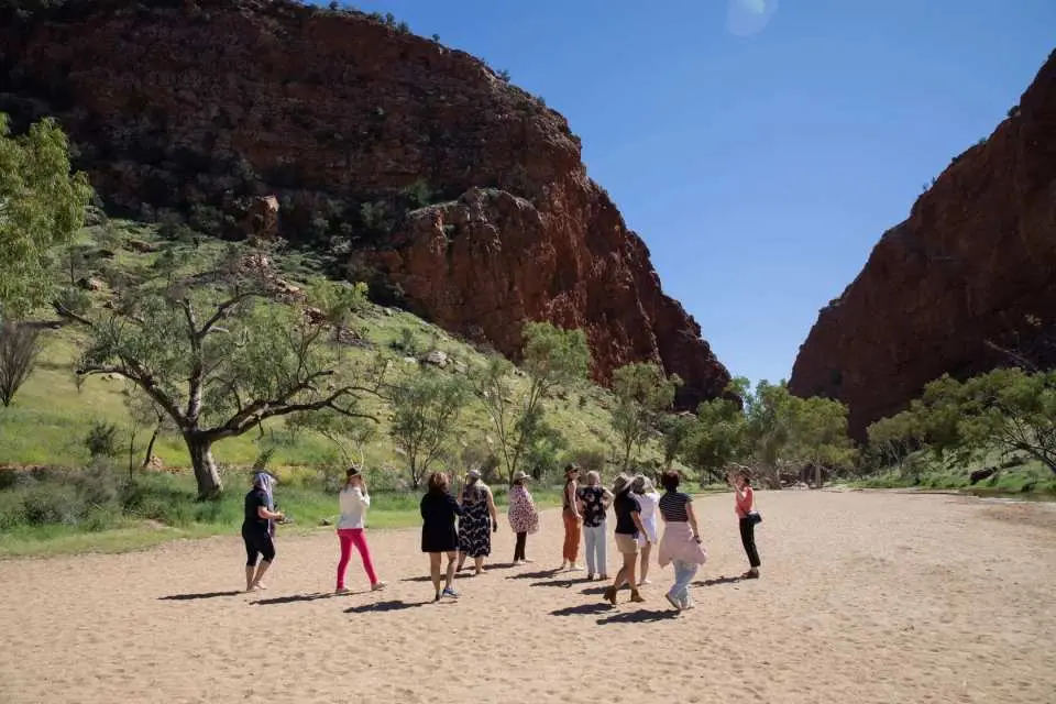 A small group gathered in a wide, sandy valley, surrounded by impressive red rock cliffs and scattered trees, enjoying a guided tour in the West MacDonnell Ranges.