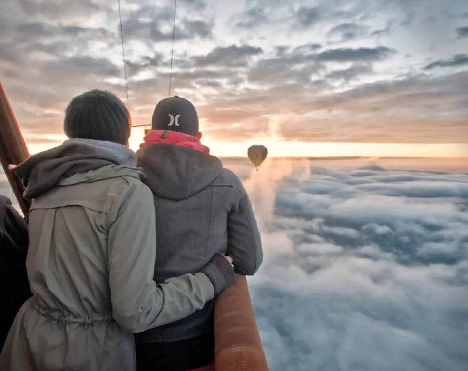 A couple embracing in a Geelong hot air balloon, gazing at the majestic sunrise over a sea of clouds, with another balloon in the distance, creating a romantic moment.