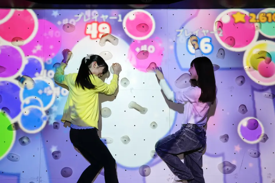 Two girls engaged in an interactive rock climbing challenge at RED° TOKYO TOWER, with colorful bubbles and scores projected onto the wall, enjoying a thrilling physical game at Tokyo's next-generation theme park.