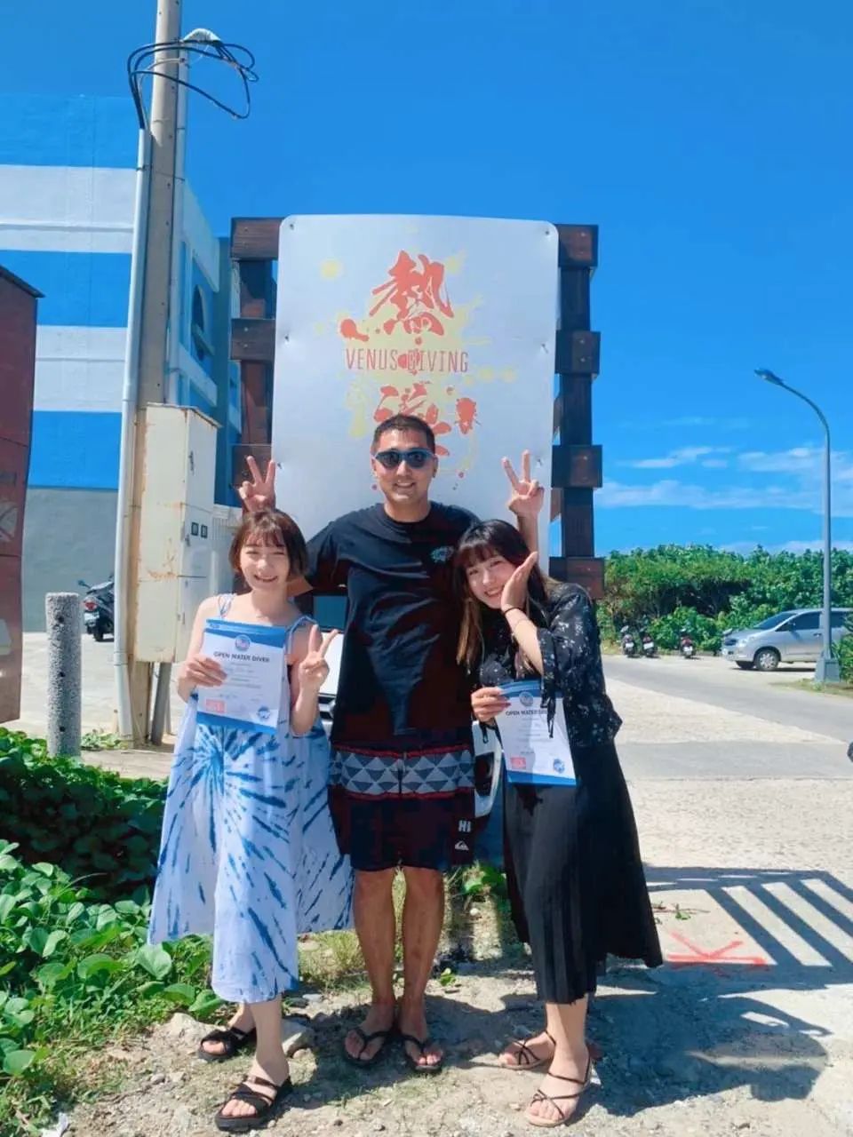 緑島の太陽の下、熱浪ダイビングセンターの看板前でダイビング学生がPADI初級オープンウォーターダイビングライセンスを手に楽しそうに記念撮影し、緑島でのダイビング体験を満喫しています。