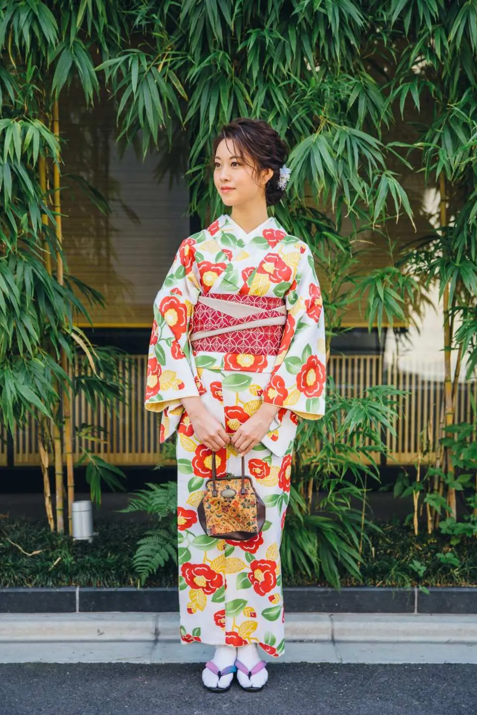 An elegant woman in a white kimono with vibrant red and orange floral patterns, holding a traditional bag, posing gracefully in front of lush green bamboo in Kyoto.