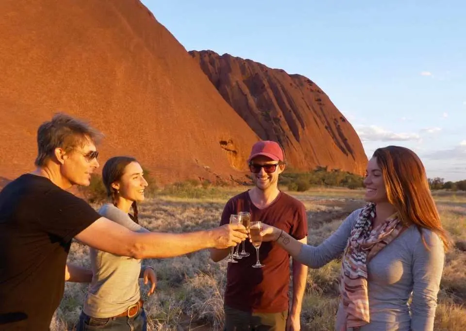 Tourists toast with sparkling wine and snacks in front of Uluru (Ayers Rock) during a magnificent sunset, celebrating an unforgettable travel experience.
