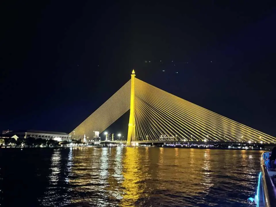Admire the dazzling night view of Rama VIII Bridge on Bangkok's Chao Phraya River from the Smile Riverside cruise, with golden lights reflecting on the water, creating a captivating scene.