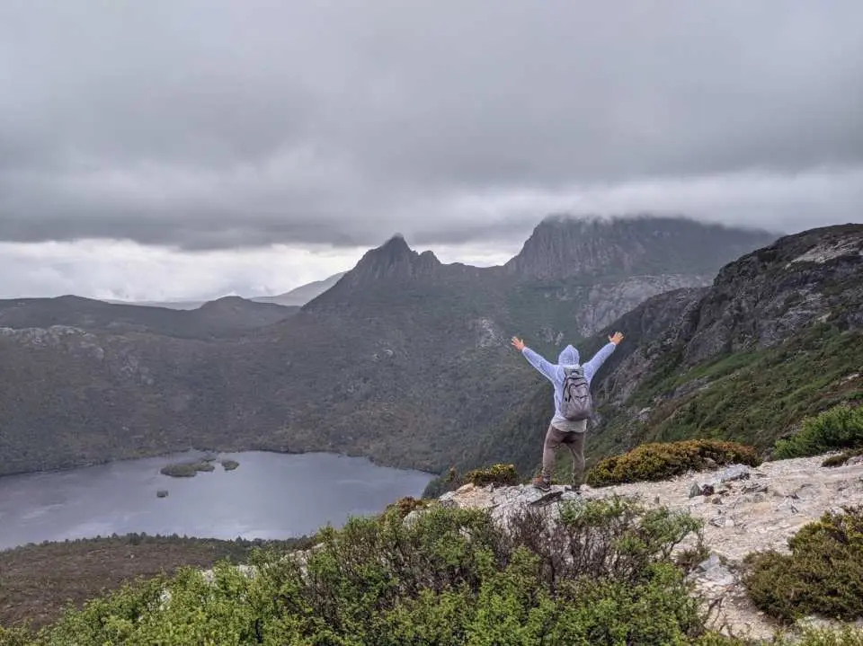 A hiker at the summit of Cradle Mountain-Lake St. Clair National Park, Tasmania, overlooking Dove Lake and rugged mountains, embracing the triumph of nature's beauty.