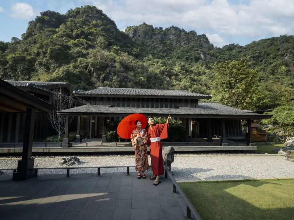 Một cặp đôi mặc kimono cầm ô đỏ, tạo dáng trong khu vườn Nhật Bản của Onsen Yokō với những ngọn núi xanh tươi phía sau, toát lên vẻ đẹp của kỳ nghỉ dưỡng.