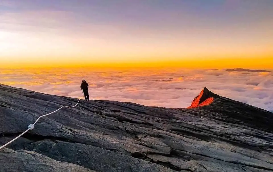 一位登山者站在京那巴魯山脊上，欣賞鐵拉達鐵索攀登途中，雲海上方絕美日出與山峰金光。