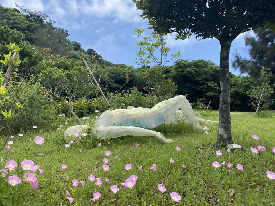 A large white human figure sculpture reclines in the lush green outdoor garden of Nanjo Museum in Okinawa, surrounded by pink flowers, embodying the museum's scene-based art and nature integration.