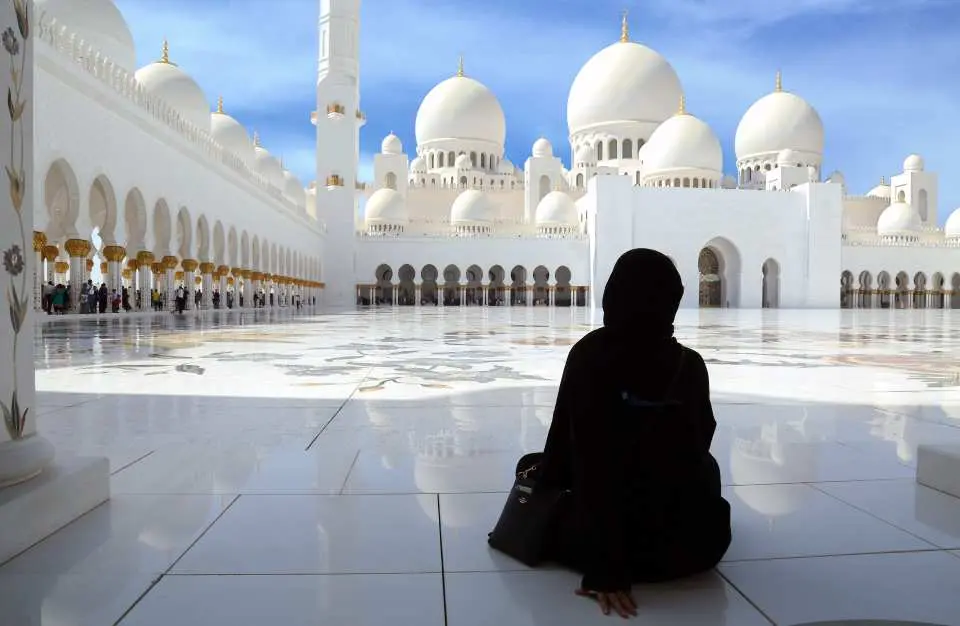 一位身穿黑色长袍的人静坐于谢赫扎耶德大清真寺广阔的白色庭院中，沉思欣赏着这座宏伟建筑的庄严与宁静。