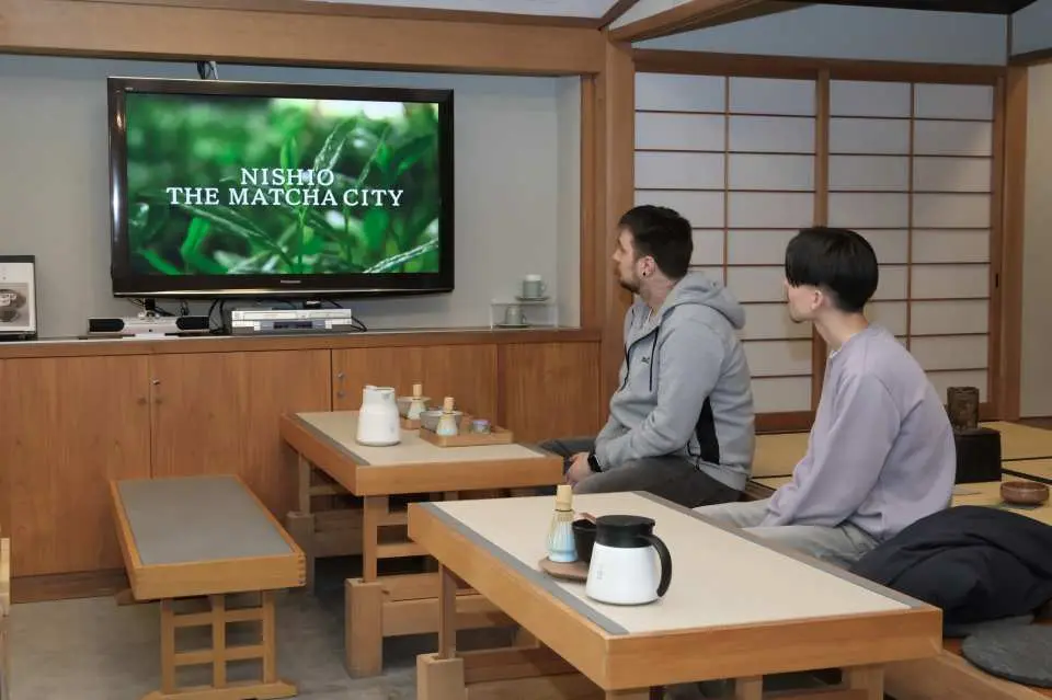 Two participants seated in a traditional Japanese setting in Nishio, watching an introductory video titled 'Nishio The Matcha City' to learn about matcha history and culture.