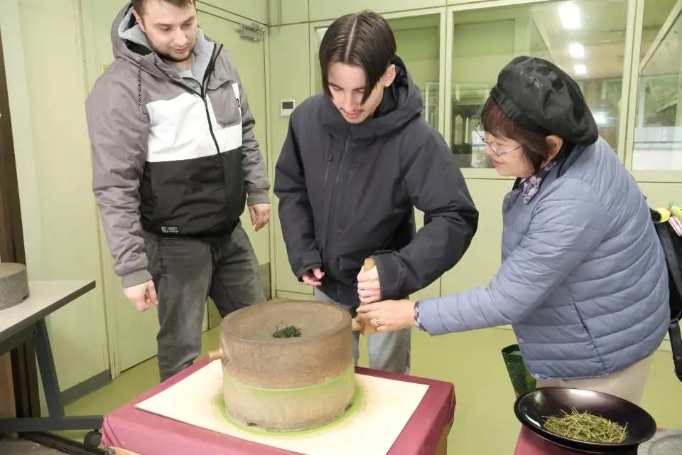 A knowledgeable guide demonstrates the traditional stone mill process of grinding tea leaves into matcha powder to two curious visitors at Aoi Seicha tea factory in Nishio.