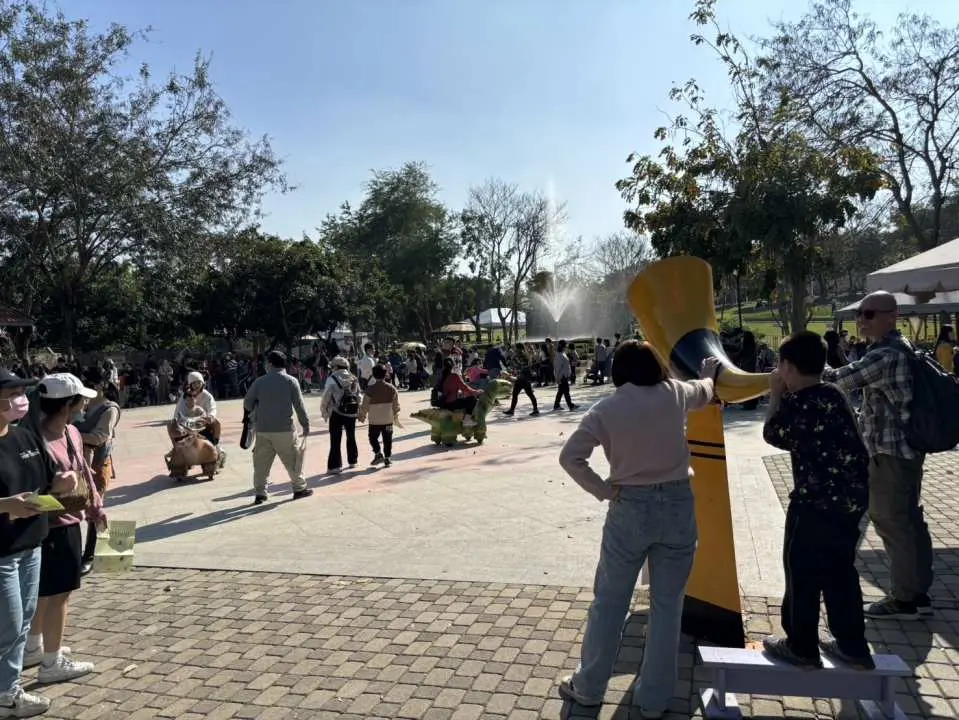 彰化古生物奇幻樂園的戶外廣場，人群聚集享受假日時光，背景是多樣化的遊樂設施，包括可能是空中叢林騎士的部分。