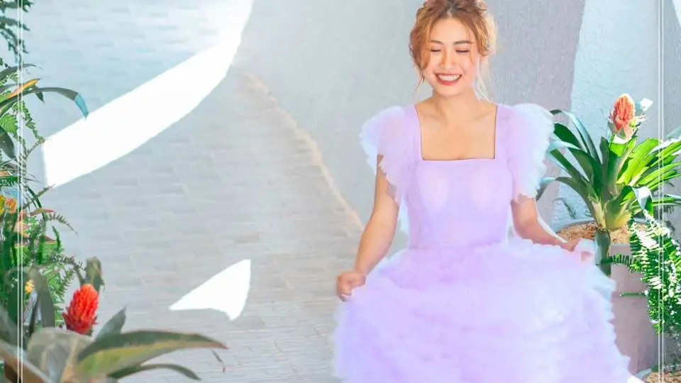 달랏 프레쉬 가든의 햇살 가득한 흰색 산책로를 연보라색 드레스를 입은 한 여성이 미소를 지으며 걷고 있습니다. 주변의 푸른 식물들 속에서 즐거움을 만끽합니다.