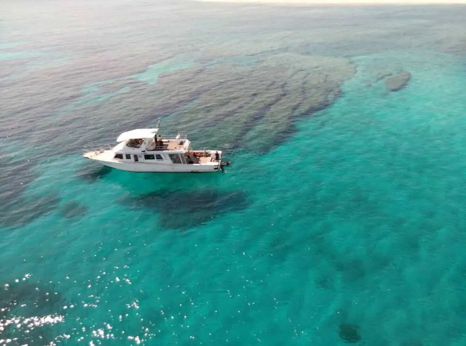 An aerial view shows a boat anchored in the pristine, shallow turquoise waters of Okinawa's Kerama Islands, revealing vibrant coral reefs below.