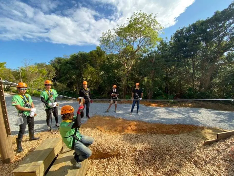 Một du khách tại điểm bắt đầu hoặc kết thúc của đường zipline trong Rừng Yanbaru ở Okinawa, đang chuẩn bị hoặc đã hoàn thành một chuyến đi đầy phấn khích, với những người tham gia khác đang quan sát.