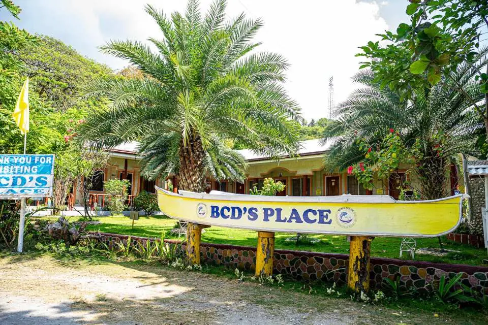 The welcoming yellow boat-shaped sign for 'BCD's Place', a facility for guests on the whale shark and Sumilon Island tour in Oslob, Cebu, surrounded by tropical foliage.