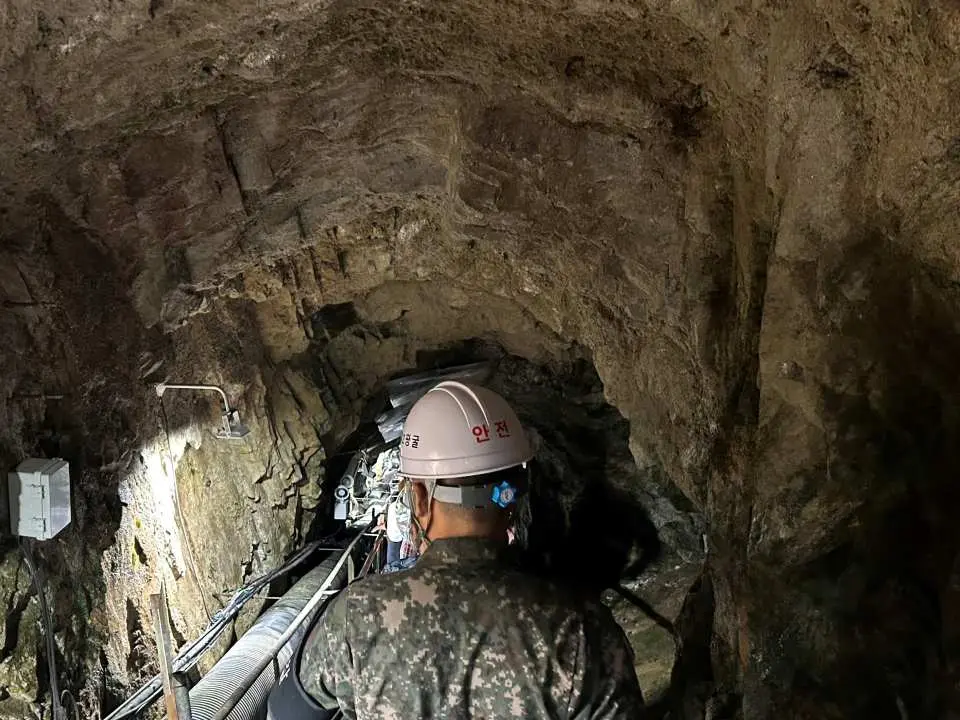 A tourist in a camouflage jacket and hard hat walks through the dark, rugged interior of the DMZ's Second Tunnel, exploring the underground passage.