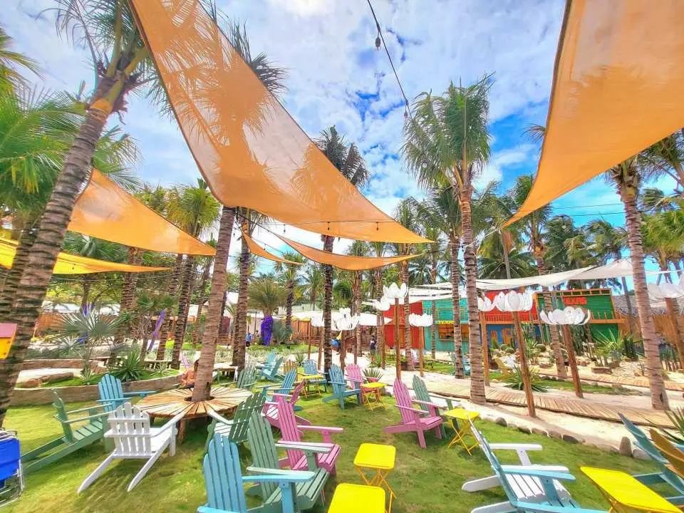 A vibrant outdoor lounge area at Mango Beach, Phan Thiet, featuring colorful chairs, small tables, and large orange sunshades amidst lush greenery and palm trees.