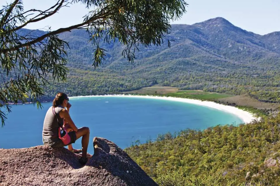 Traveler sits high above Wineglass Bay, Tasmania, admiring the magnificent crescent-shaped beach and sapphire blue bay surrounded by lush green mountains.