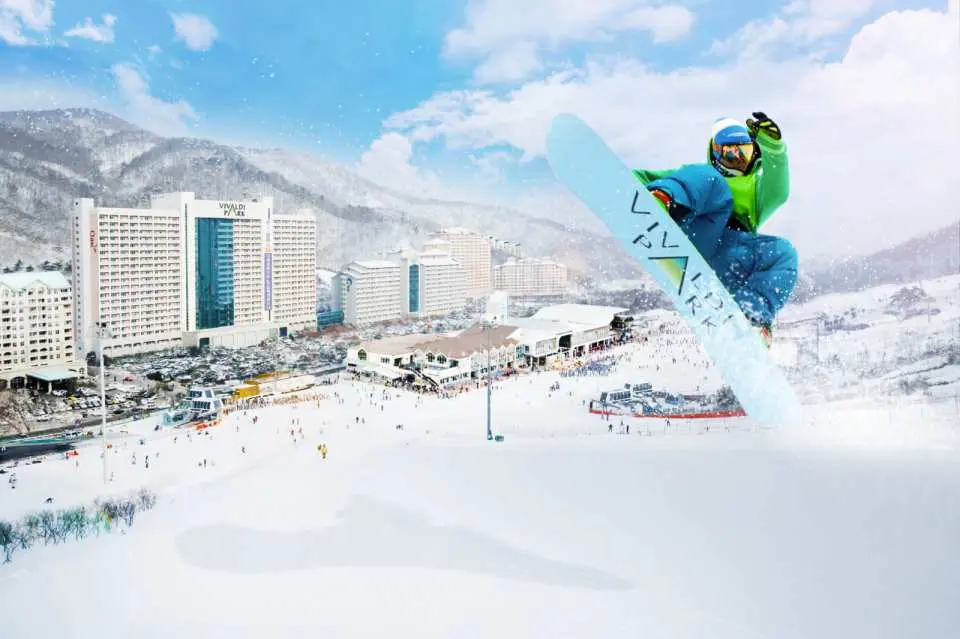 นักสโนว์บอร์ดกระโดดลอยตัวกลางอากาศเหนือลานสกี Vivaldi Park Ski Resort โดยมีอาคารรีสอร์ตและภูเขาที่ปกคลุมด้วยหิมะเป็นฉากหลัง แสดงให้เห็นถึงความตื่นเต้นของกีฬาฤดูหนาวสุดขีด.