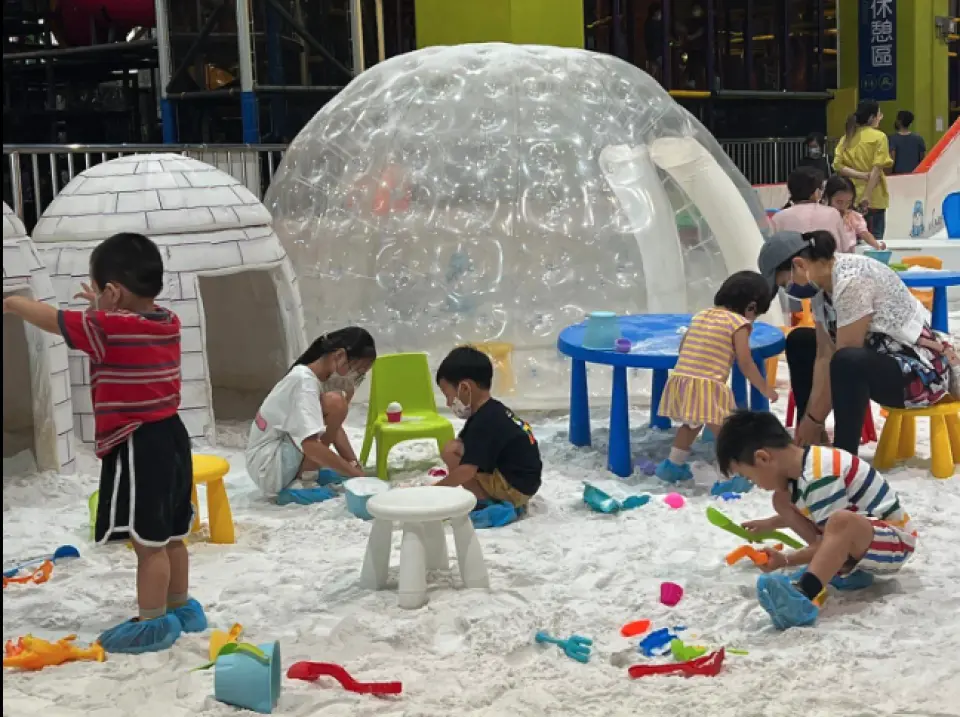 Children in blue snow booties happily build snowmen and play with snow in the Shang Shun World Snow Paradise children's play area, featuring a charming igloo and an inflatable bubble tent.