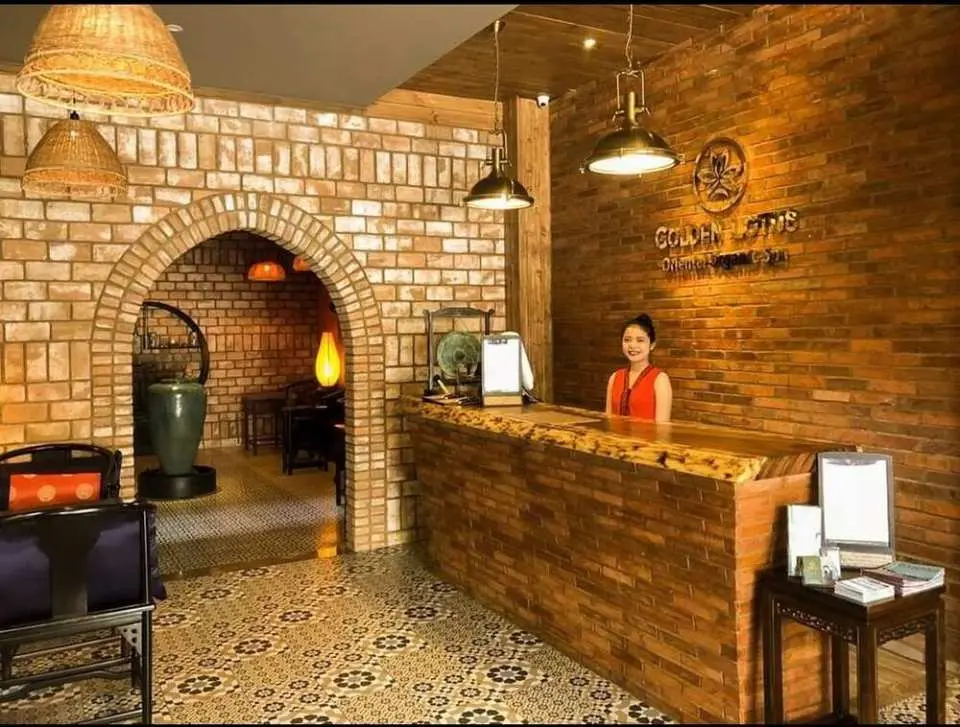 다낭 골든 로터스 오리엔탈 오가닉 스파의 환영하는 리셉션 공간. 미소 짓는 직원이 전통 벽돌 장식 속에서 손님들을 맞이할 준비가 되어 있습니다.