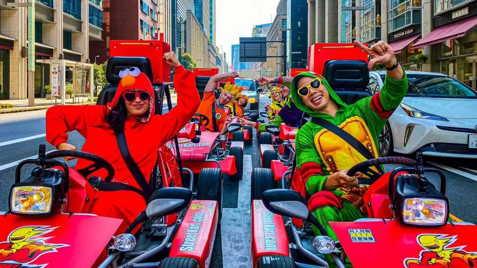 Tourists in vibrant cartoon costumes happily drive street karts through Tokyo's city streets, posing with smiles against a backdrop of modern skyscrapers, enjoying a fun and unique experience.