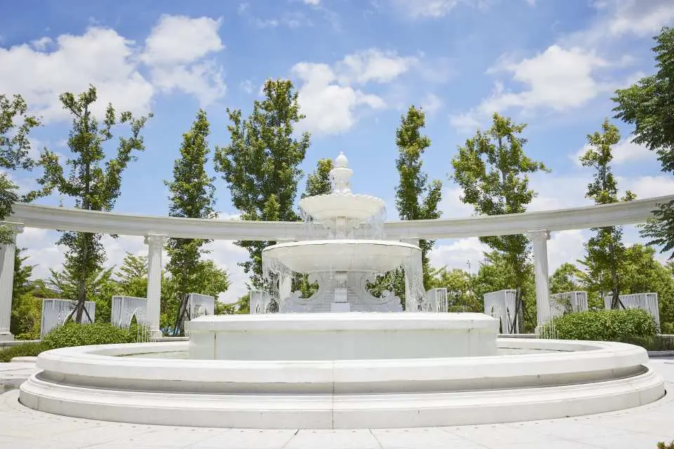 嘉義蓋婭莊園戶外庭園的白色三層噴水池，周圍綠樹環繞，古典羅馬柱拱門在背景若隱若現，是悠閒的休憩場所。