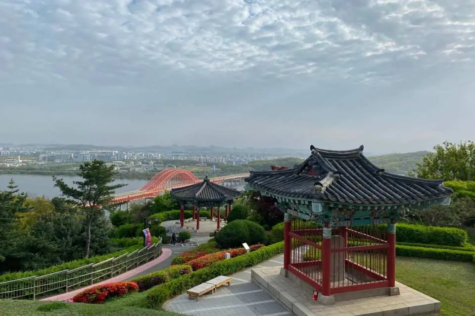 從幸州山城鳥瞰漢江壯麗景色，傳統亭閣與綠意盎然的山坡點綴其中，遠方紅色橋樑與城市天際線清晰可見。