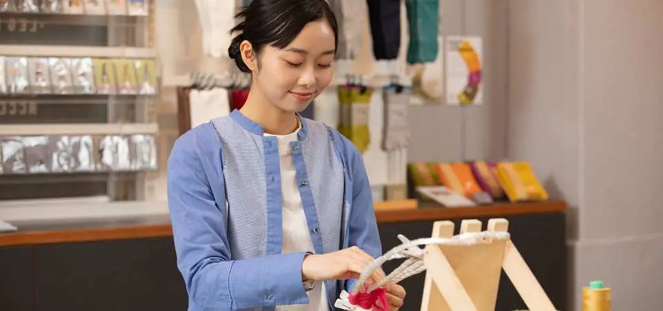 A young woman intently weaving fabric sandals at a workshop in Ryogoku, Tokyo, with colorful yarns and fabrics in the background, showcasing the joy of craftsmanship.