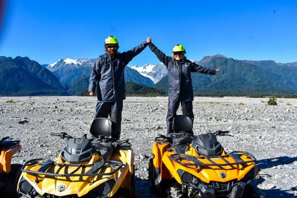 Two adventurers triumphantly stand on yellow quad bikes in New Zealand's Franz Josef Glacier riverbed, with majestic mountains under a clear blue sky.