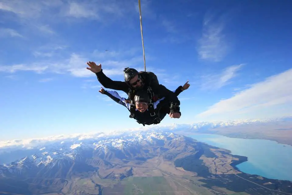 Two tandem skydivers in exhilarating freefall above Mount Cook, smiling with arms outstretched, overlooking New Zealand's vibrant turquoise Lake Pukaki and majestic snow-capped mountains.