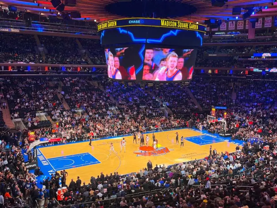 NBA New York Knicks basketball game at Madison Square Garden, with a packed crowd immersed in the action, a giant screen providing an optimal viewing experience, and a vibrant atmosphere.