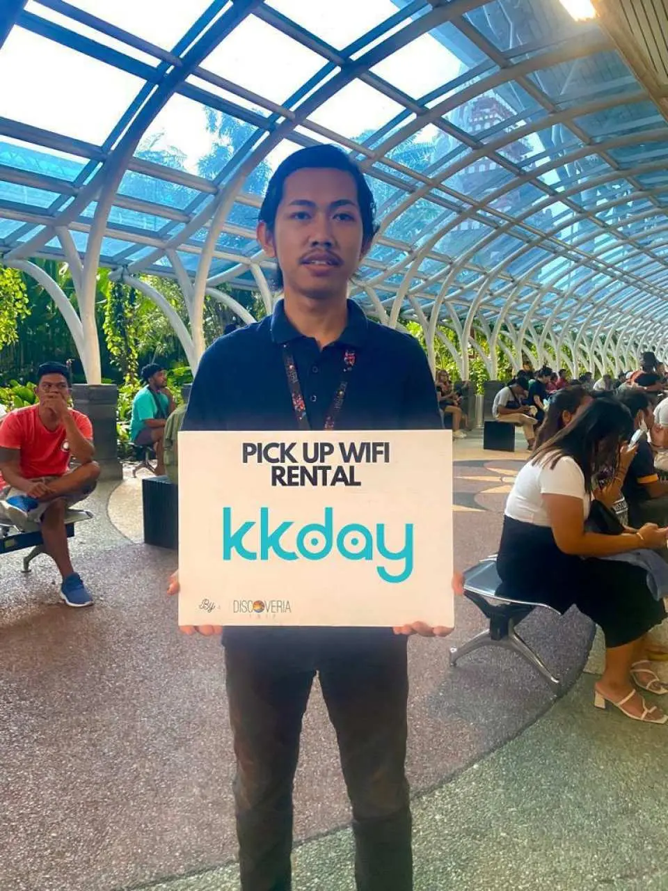 バリ島空港の指定された受け取り地点で「PICK UP WIFI RENTAL kkday」と書かれた看板を持つKKdayスタッフ。旅行者に4GポータブルWi-Fiルーターの受け取りサービスを案内しています。