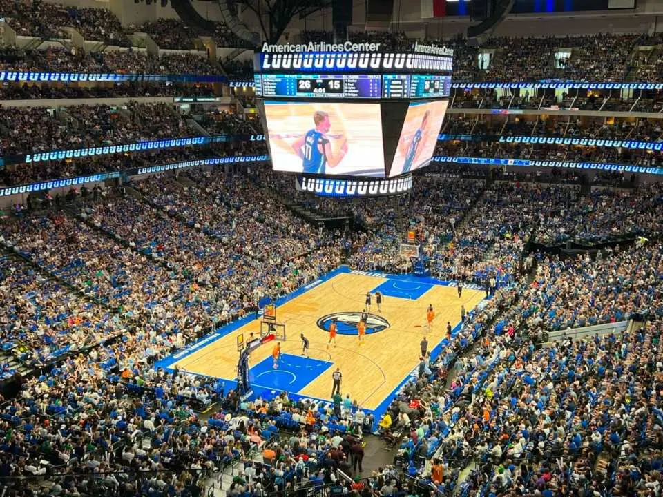 Overhead view of a bustling NBA basketball game at Dallas' American Airlines Center, with a packed arena and a giant scoreboard displaying thrilling match highlights, creating an immersive experience.